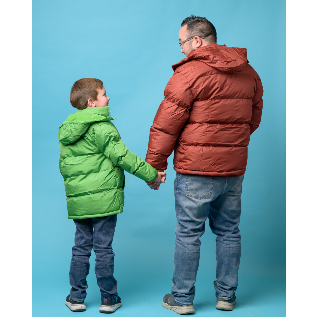 Man and child holding hands wearing wholesale bulk puffer coats/jackets against a blue background